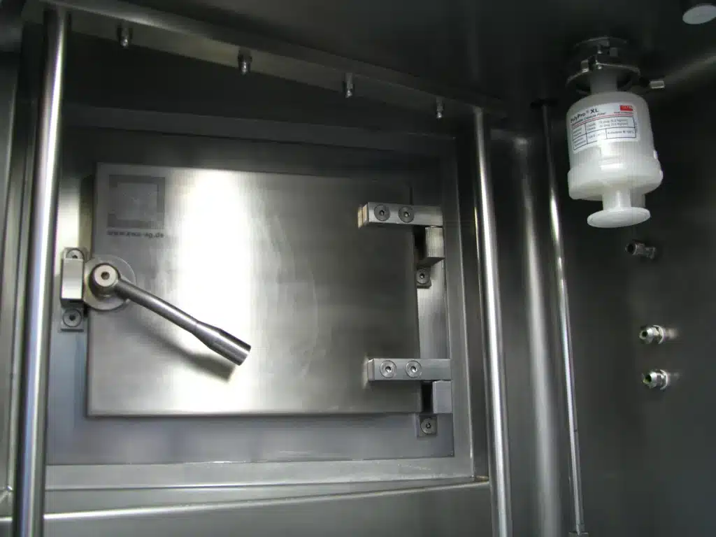 Edelstahltür mit Griff eines EWA VETK 8, eingebaut in die Wand eines Isolators. Rechts daneben sind ein weißer Filter und zwei Anschlüsse zu sehen.